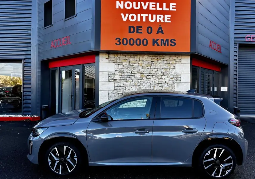 Profil côté gauche d'une Peugeot 208 Hybrid 136 eDCS6 GT gris foncé stationnée devant un bâtiment avec panneau orange.