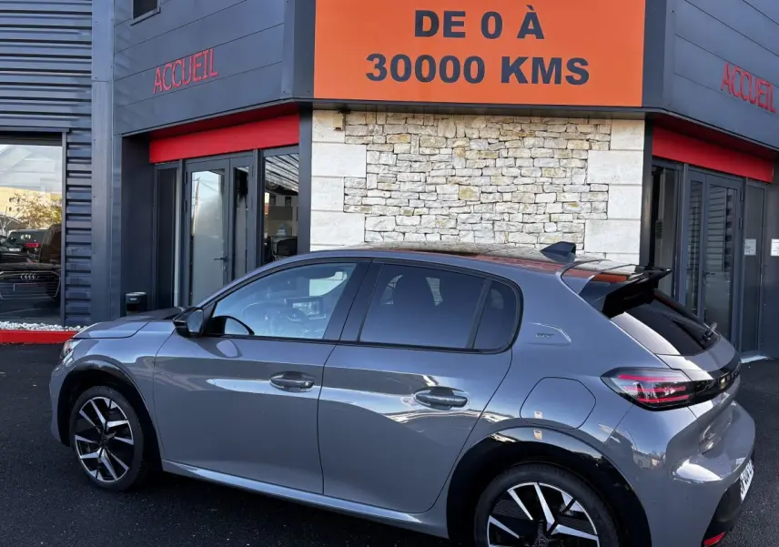 Peugeot 208 Hybrid 136 eDCS6 GT gris foncé vue de profil côté gauche devant un bâtiment avec panneau orange.