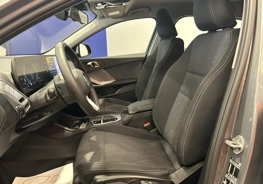 Vue intérieure côté conducteur de la BMW Série 1 120DA 2024, sièges tissu gris et tableau de bord moderne.