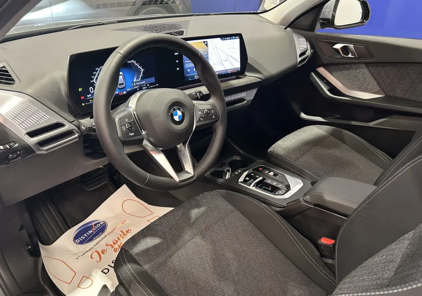 Intérieur de la BMW Série 1 120DA 2024, vue côté conducteur, tableau de bord moderne avec écran digital et sièges tissu gris.