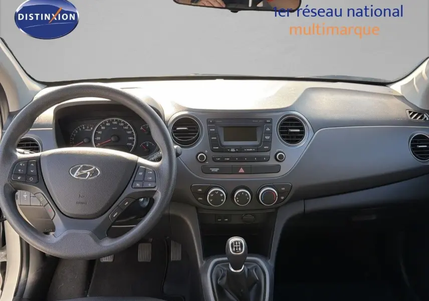 Vue intérieure du tableau de bord et volant noir d'une Hyundai i10 blanche 2018 avec boîte manuelle et commandes au volant.