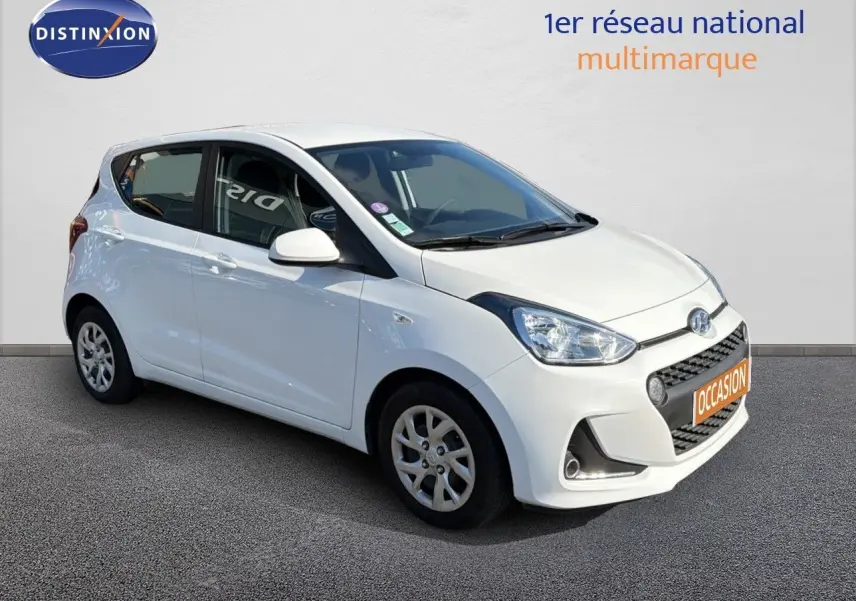 Hyundai i10 blanche vue 3/4 avant droit, compacte cinq portes avec jantes alliage et calandre noire distinctive.