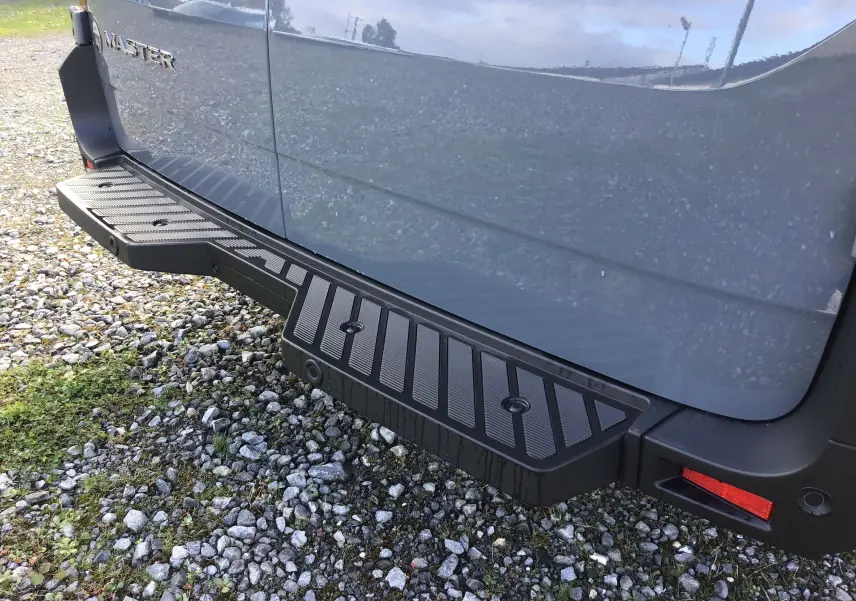 Gros plan sur le marchepied arrière noir du Renault Master gris urbain, vue latérale côté droit sur sol gravillonné.