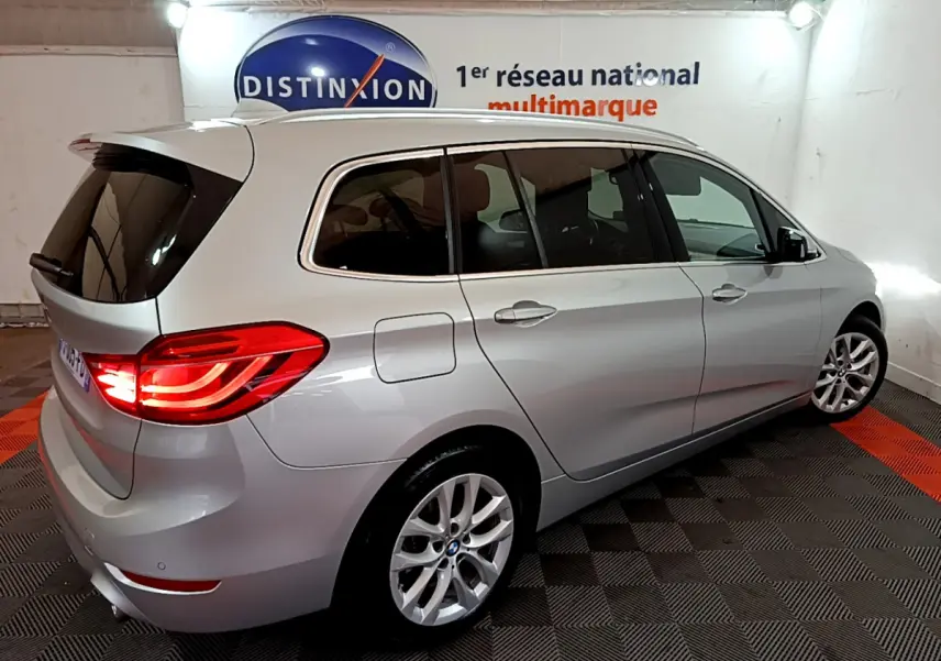 BMW Série 2 Gran Tourer 218d gris clair vue 3/4 arrière droit avec feux arrière allumés en intérieur showroom.