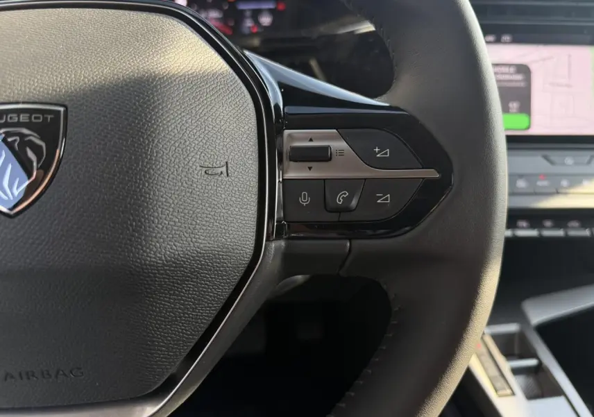 Gros plan sur le volant cuir noir de la Peugeot 308 2025, avec commandes multimédia et logo Peugeot visible.