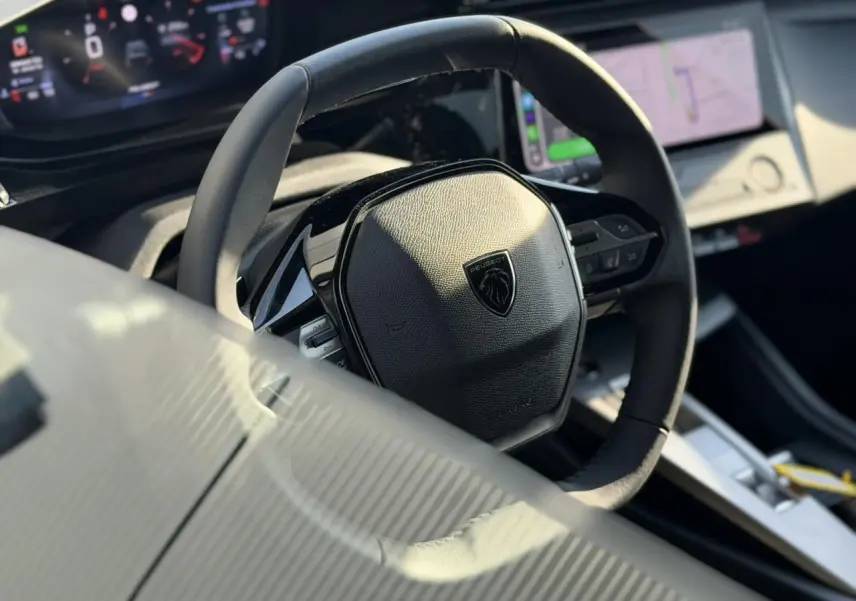Gros plan sur le volant cuir noir de la Peugeot 308 2025, avec tableau de bord numérique et écran tactile en arrière-plan.