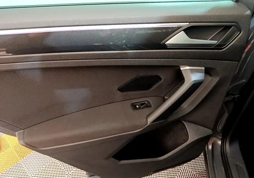 Vue intérieure côté droit de la porte du Volkswagen Tiguan gris clair, avec poignée argentée et panneau noir texturé.