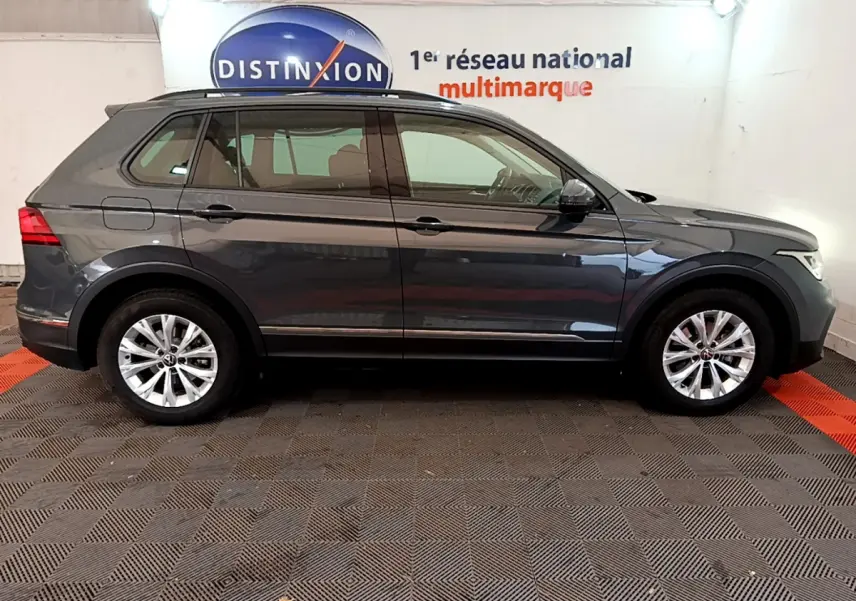 Volkswagen Tiguan gris clair vu de profil côté gauche dans un showroom avec logo Distinxion en arrière-plan.
