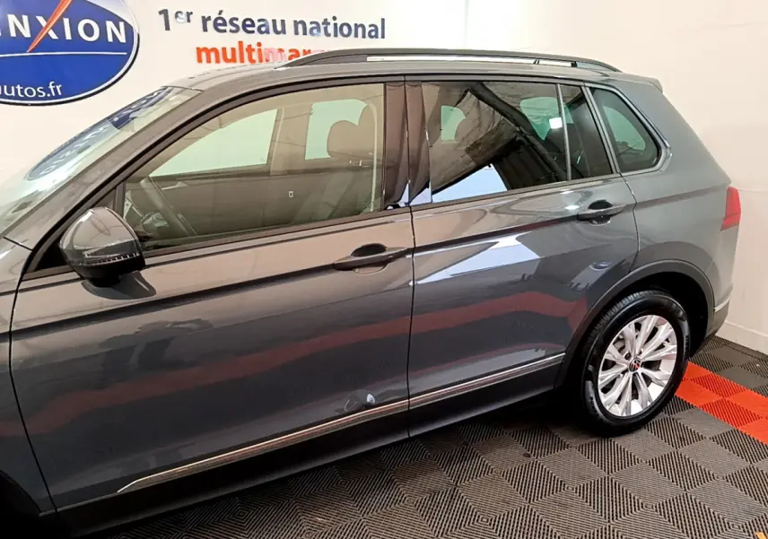 Vue latérale côté gauche d'un Volkswagen Tiguan gris clair 2021, avec jantes alliage et barres de toit noires visibles.