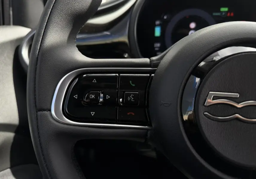 Gros plan sur le volant noir Soft-Touch de la Fiat 500 électrique 2024, avec commandes intégrées et logo 500 au centre.