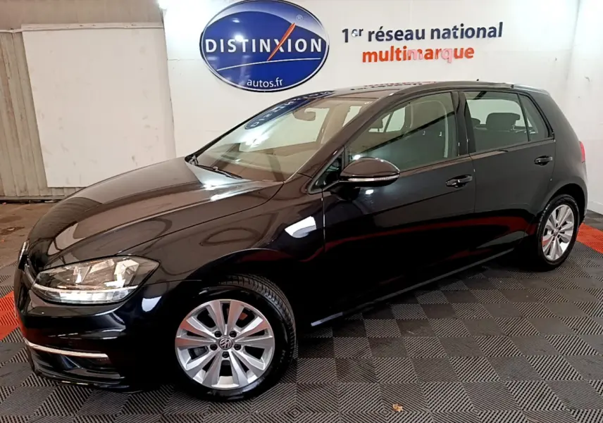 Volkswagen Golf noire vue en 3/4 avant droit, avec jantes alliage et phares allumés en intérieur showroom.