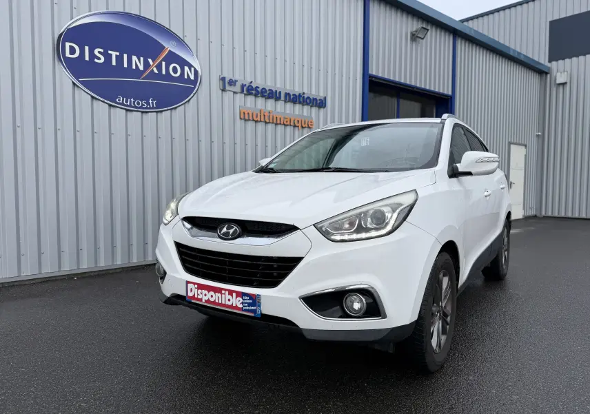 Hyundai ix35 blanc vue 3/4 avant droit, avec phares allumés et plaque "Disponible" devant un bâtiment industriel.