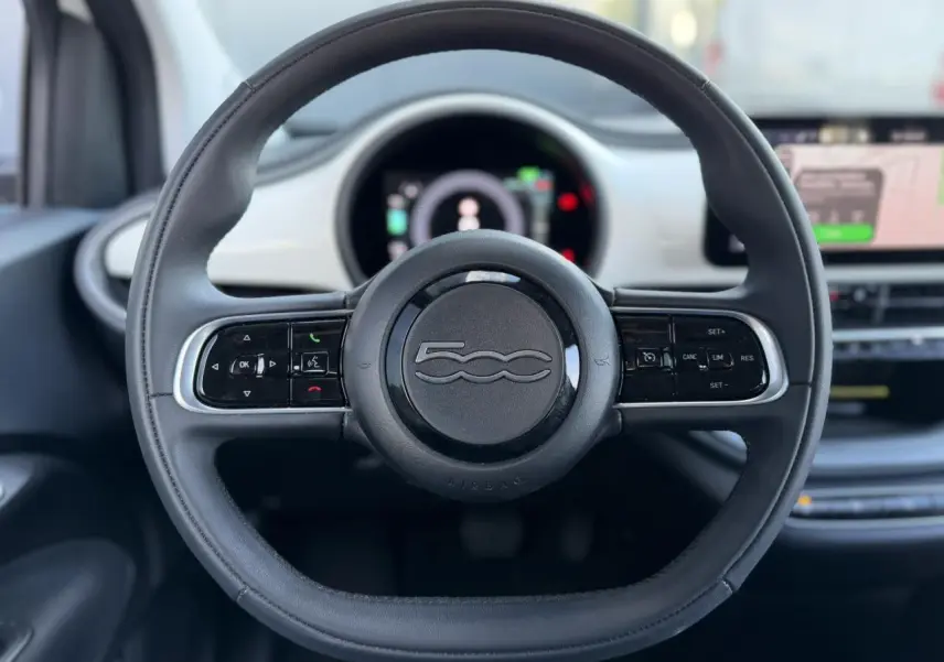 Volant noir Soft-Touch de la Fiat 500 électrique 2024, avec commandes intégrées et tableau de bord numérique en arrière-plan.