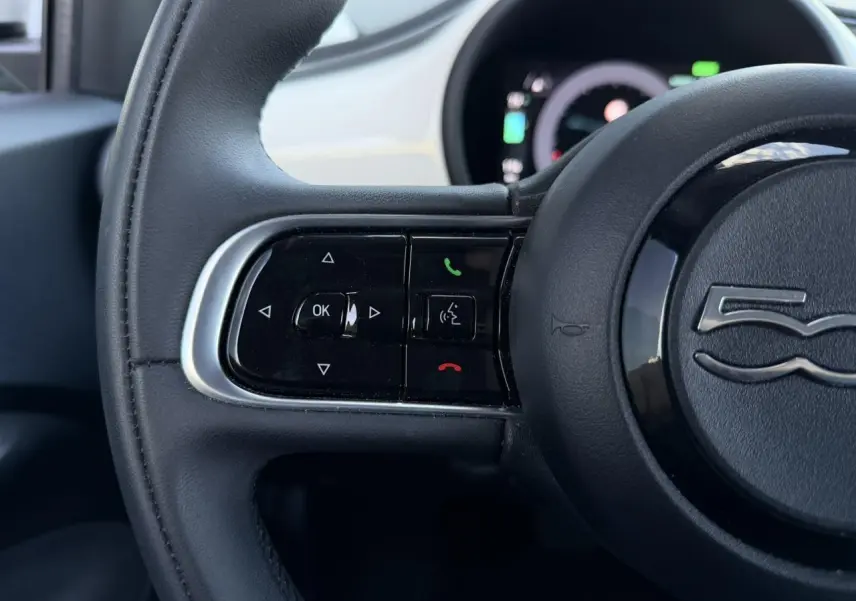 Gros plan sur la partie gauche du volant noir de la Fiat 500 électrique 2024 avec commandes intégrées et logo central.