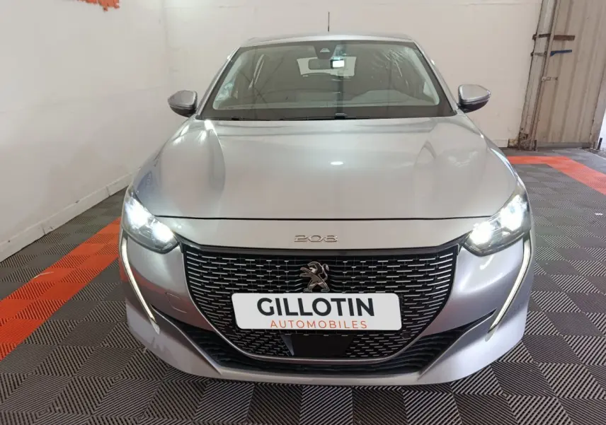 Vue frontale d'une Peugeot 208 électrique gris clair avec phares LED allumés et calandre noire distinctive.