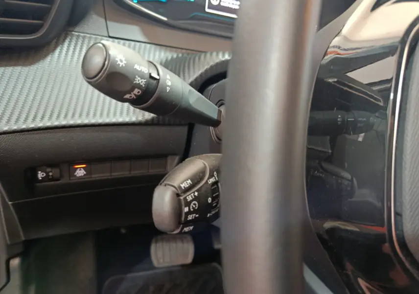 Gros plan sur la colonne de direction et commandes au volant d'une Peugeot 208 électrique gris clair, tableau de bord partiellement visible.