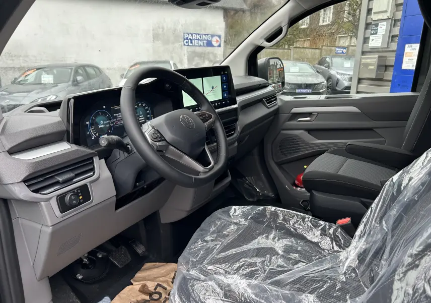 Vue intérieure côté conducteur du Volkswagen Transporter T7 ProCab 2026, montrant le tableau de bord numérique et les sièges en tissu protégés par film plastique.