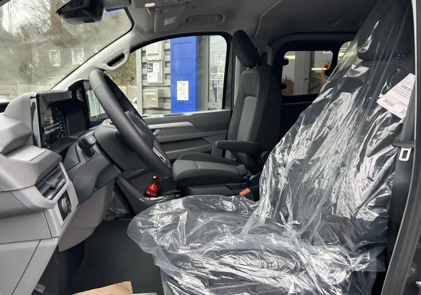 Vue intérieure côté conducteur du Volkswagen Transporter T7 ProCab, sièges noirs avec protection plastique, tableau de bord moderne.