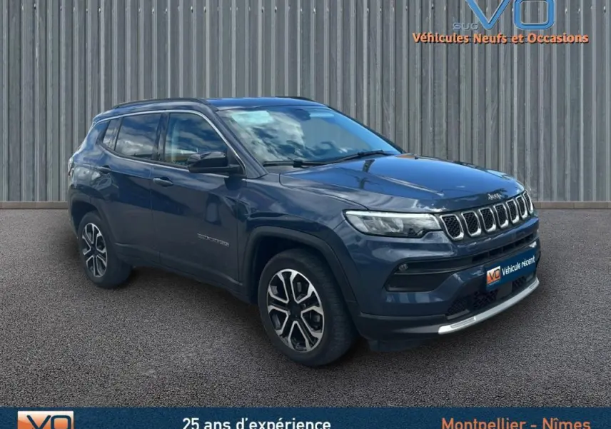 Jeep Compass bleu 3/4 avant droit avec barres de toit et jantes 18 pouces, stationné devant un mur métallique.