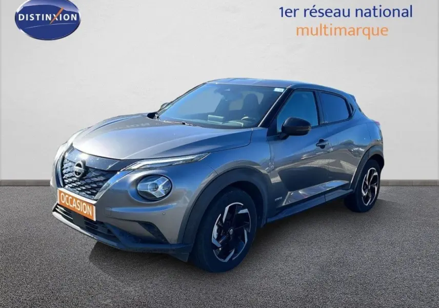 Nissan Juke Hybrid gris squale métal en 3/4 avant droit, avec calandre noire et phares ronds distinctifs.