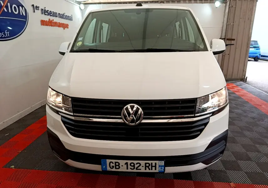 Vue avant d’un Volkswagen Transporter blanc 2021 avec calandre noire et phares allumés dans un garage.
