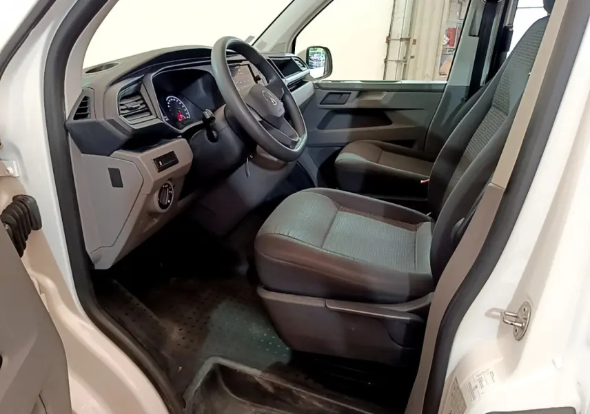 Vue intérieure côté conducteur du Volkswagen Transporter blanc, montrant le tableau de bord et les sièges avant gris.