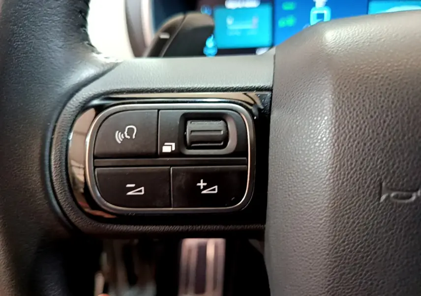 Gros plan sur les commandes gauche du volant cuir noir du Citroën C5 Aircross Hybrid 225 ë-EAT8 Shine, avec écran digital flou en arrière-plan.