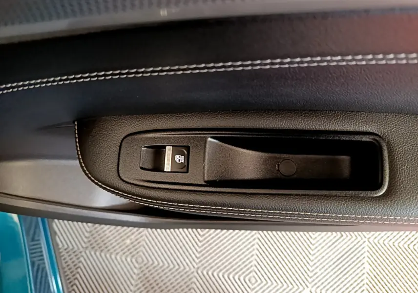 Gros plan sur la poignée intérieure et commande de vitre du côté gauche, avec surpiqûres blanches et teinte bleu visible à l’extérieur.