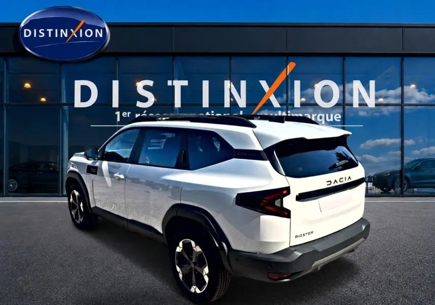 Vue 3/4 arrière droite du Dacia Bigster blanc avec barres de toit et jantes alu devant un showroom Distinxion.
