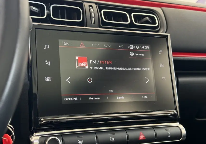 Écran tactile central de la Citroën C3 noire, vue rapprochée du tableau de bord avec commandes climatisation et radio FM affichée.