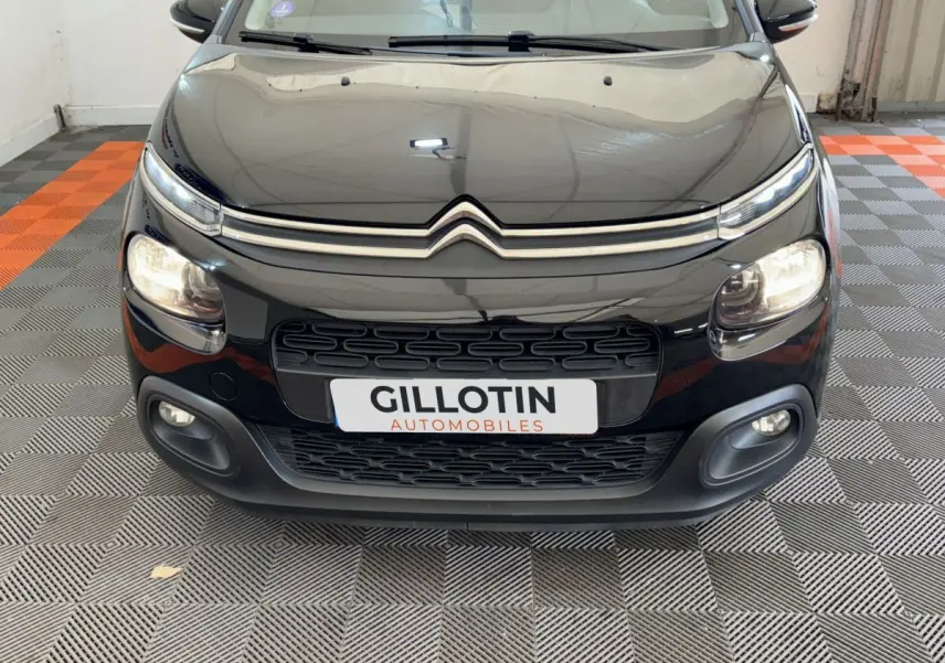 Vue frontale rapprochée d'une Citroën C3 noire avec phares allumés et calandre distinctive en intérieur showroom.