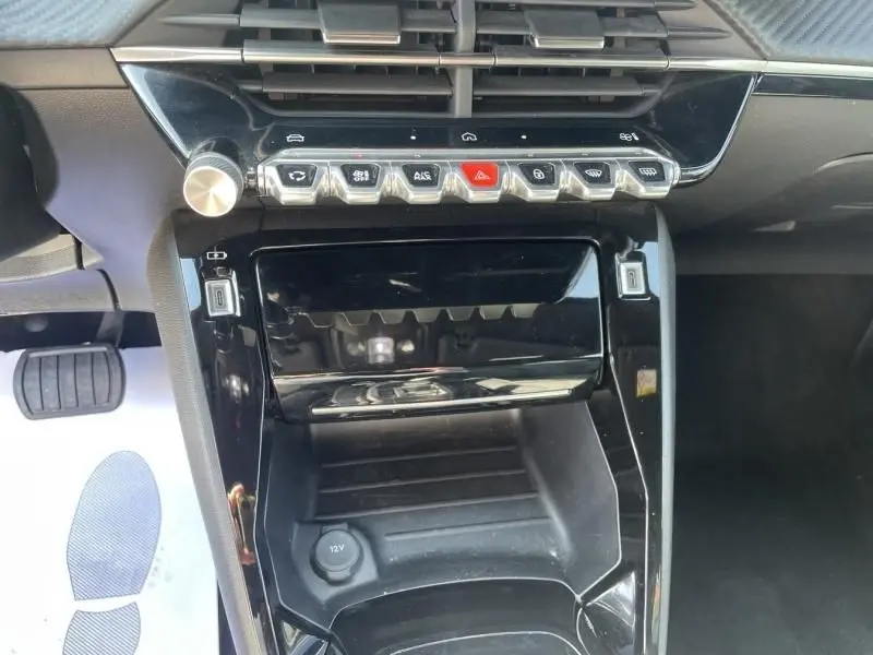 Vue rapprochée de la console centrale noire brillante du Peugeot 2008 Hybrid 2025 avec commandes tactiles et prise 12V.