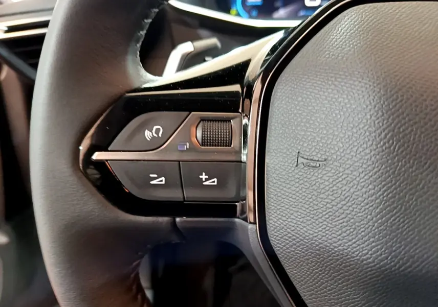 Gros plan sur le volant cuir noir de la Peugeot 208 2022, avec commandes audio et tableau de bord numérique en arrière-plan.