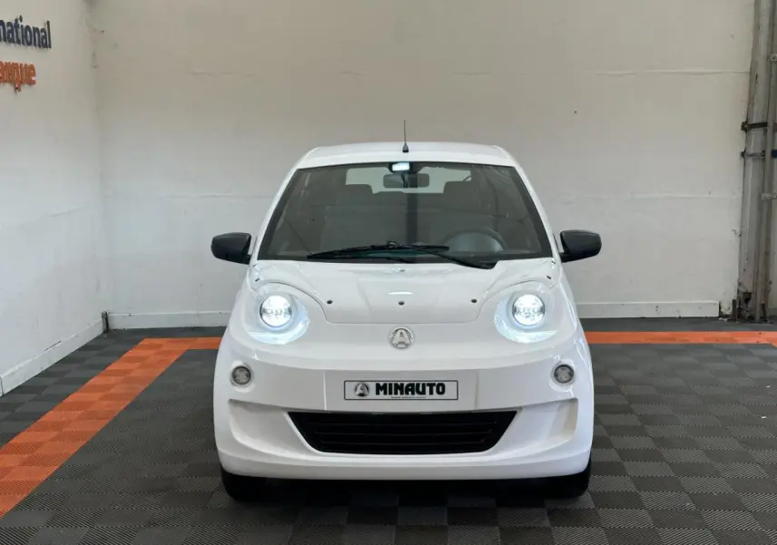 Vue frontale d'une AIXAM MINAUTO E ACCESS blanche 2025 avec phares allumés et rétroviseurs noirs côté conducteur.