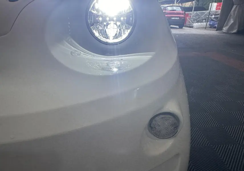 Gros plan sur le phare avant droit allumé d'une Aixam Minauto E Access blanche 2025 avec feu antibrouillard visible.