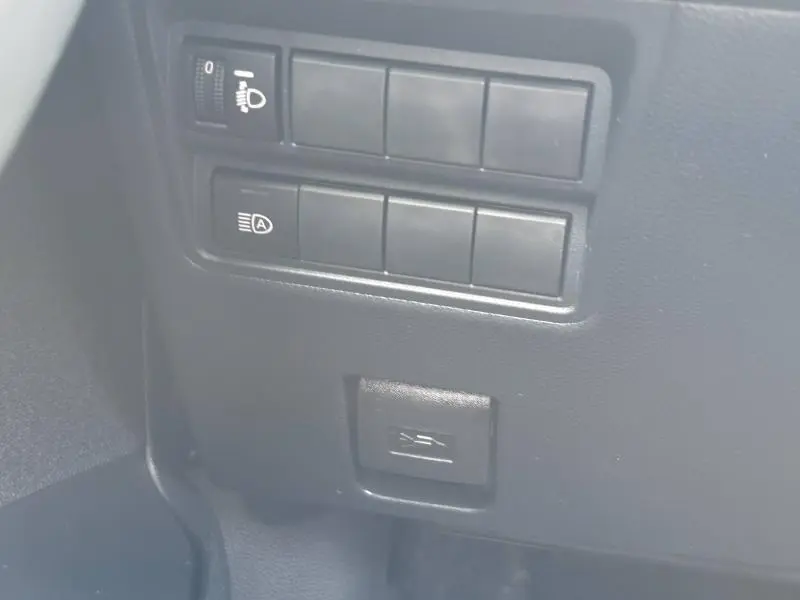 Gros plan sur les commandes de réglage des phares et bouton d'assistance sur le tableau de bord noir de la Toyota Yaris 116h Dynamic blanche.