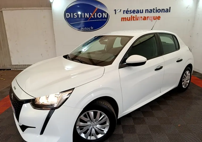Peugeot 208 blanc vue 3/4 avant droit en intérieur, avec phares allumés et jantes alliage visibles.