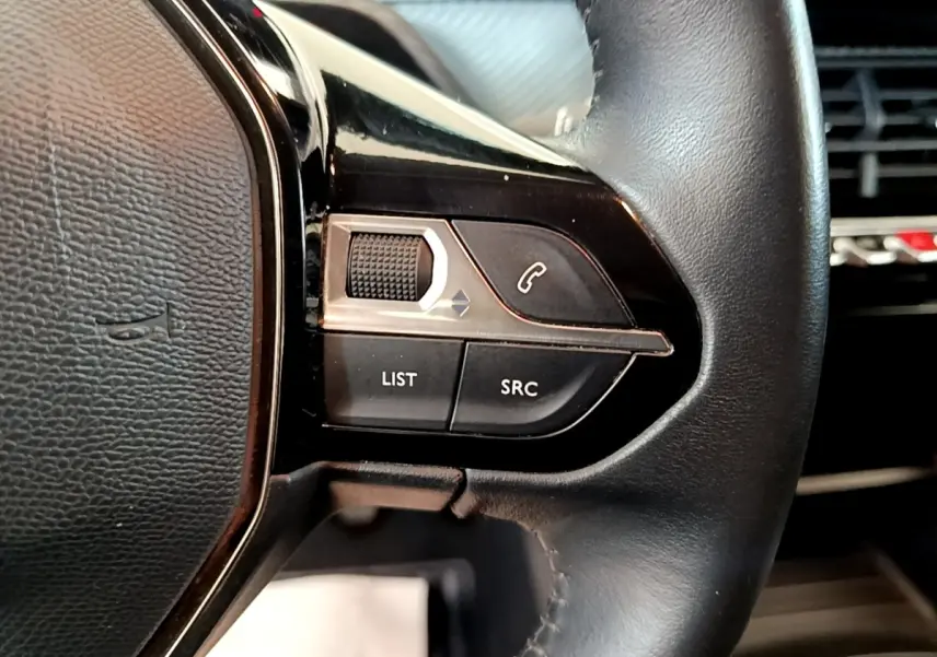 Gros plan sur les commandes gauche du volant en cuir noir d'une Peugeot 208 blanche, avec boutons de téléphone et liste.