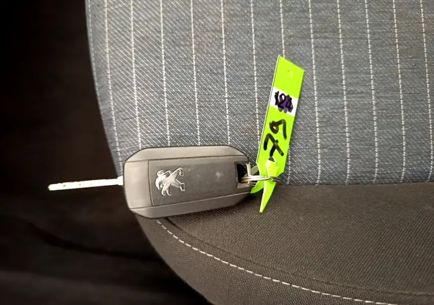 Clé de voiture Peugeot posée sur un siège tissu gris à rayures blanches, avec une étiquette verte attachée.