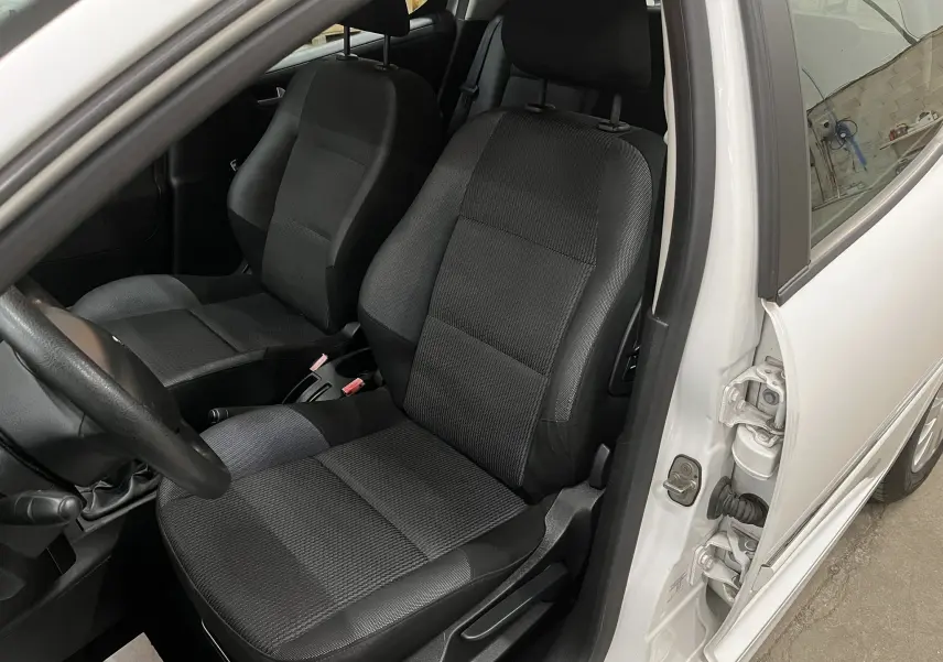 Vue intérieure côté conducteur d’une Peugeot 207 blanche de 2011, sièges tissu gris-noir et volant visible.