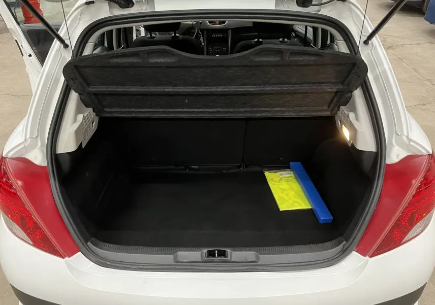 Coffre ouvert d'une Peugeot 207 blanche vue de l'arrière, avec tapis noir et gilet de sécurité visible à droite.