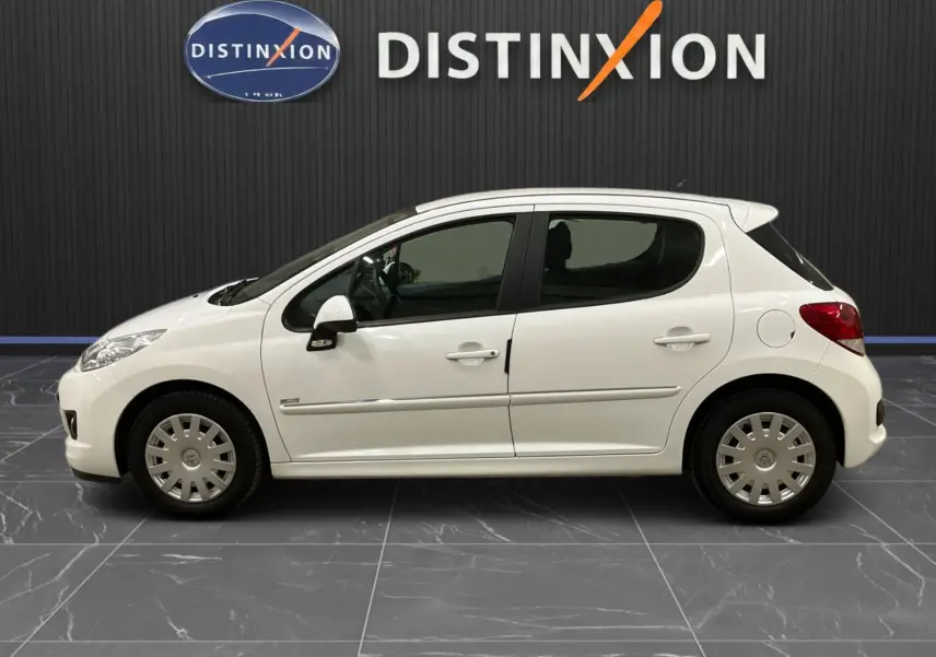 Profil droit d'une Peugeot 207 blanche 5 portes avec baguette chrome et jantes acier en intérieur showroom.