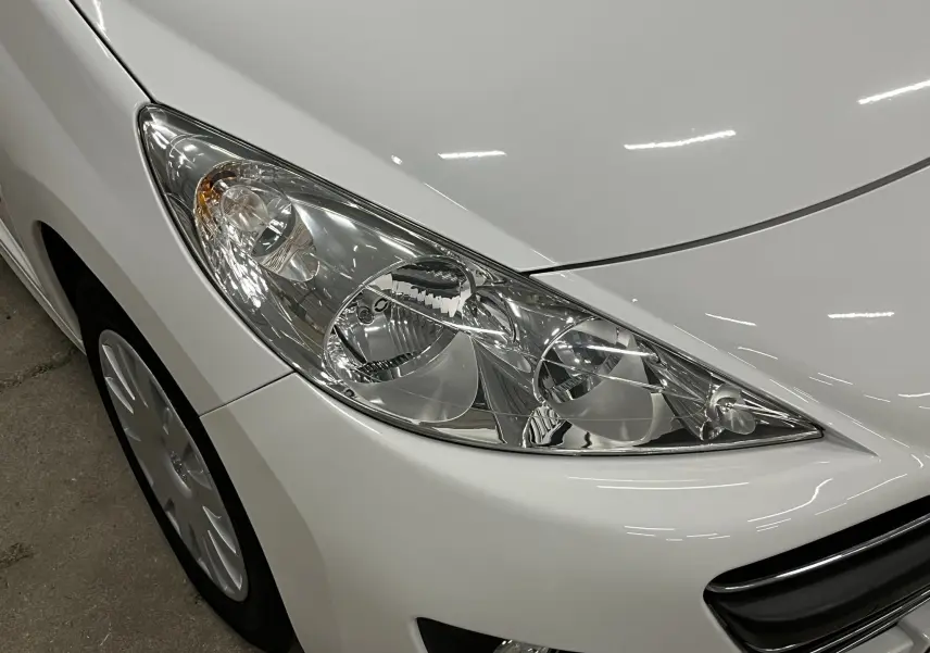 Gros plan sur le phare avant droit d'une Peugeot 207 blanche, avec calandre et jante visibles.