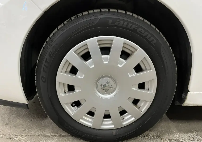 Gros plan sur la roue avant gauche d'une Peugeot 207 blanche, avec enjoliveur argenté et logo lion au centre.