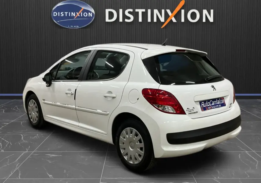 Vue 3/4 arrière droite d'une Peugeot 207 blanche avec baguettes chrome et béquet, stationnée en intérieur showroom.