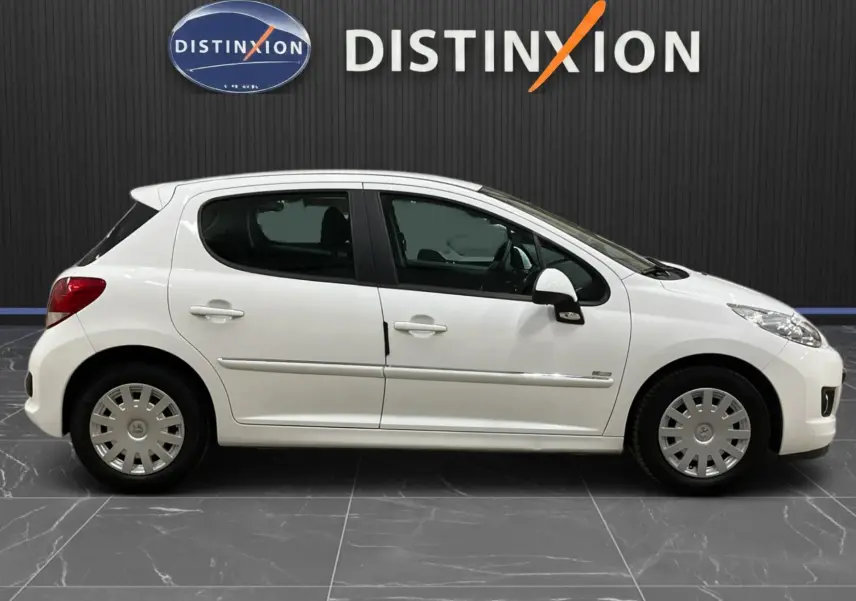 Peugeot 207 blanche vue de profil côté gauche, avec baguette de porte chromée et jantes acier à enjoliveurs.