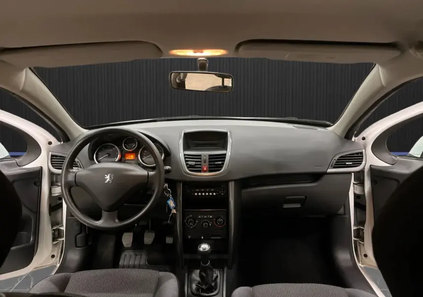 Vue intérieure frontale du tableau de bord d'une Peugeot 207 blanche de 2011 avec volant, levier de vitesse manuel et sièges tissu gris-noir.