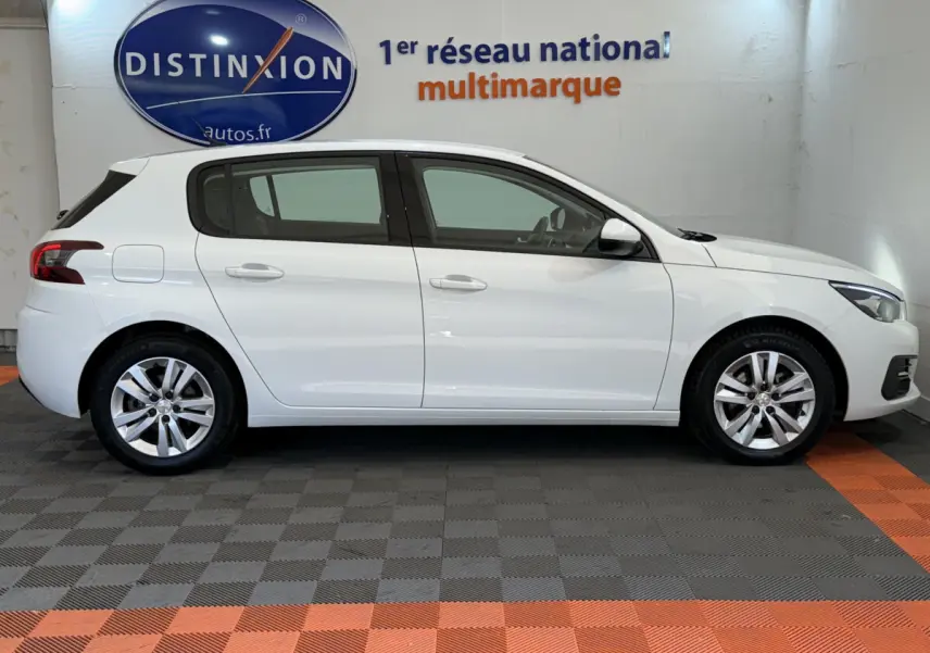 Profil côté gauche d'une Peugeot 308 blanche 2021 garée en intérieur sur sol gris et orange, devant un mur avec logo Distinxion.