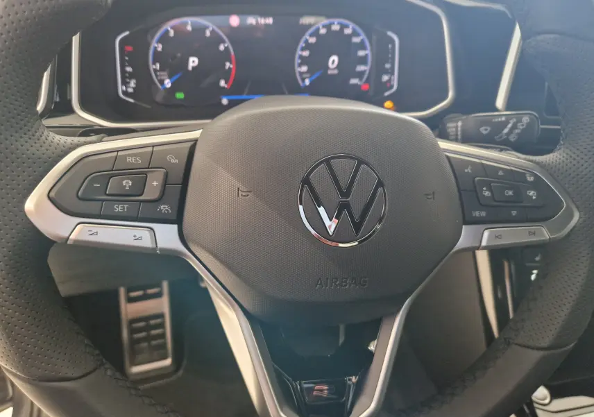 Vue rapprochée du volant multifonction noir perforé du Volkswagen T-Roc 2025 avec tableau de bord numérique en arrière-plan.