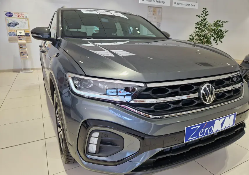 Vue 3/4 avant gauche du Volkswagen T-Roc gris Indium Metal avec calandre R-Line et plaque Zero KM en concession.